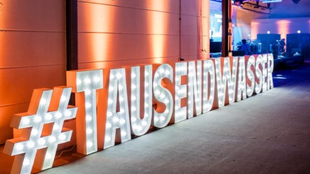 Ein beleuchtetes Schild mit dem Hashtag "#TAUSENDWASSER" steht vor einer Event-Umgebung. Die Buchstaben sind kräftig und deutlich, umgeben von orangefarbigen Lichtern.