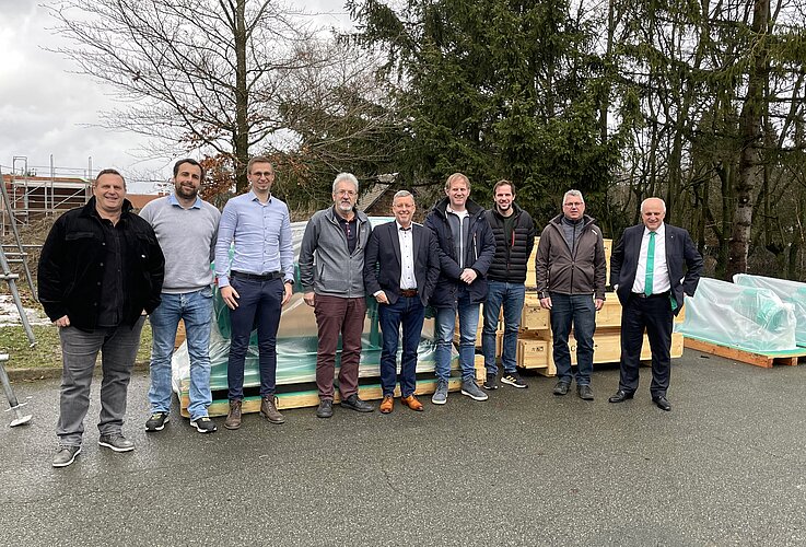 Eine Gruppe von zehn Männern steht auf einer gepflasterten Fläche, umgeben von Bäumen und Baumaterial. Sie tragen Freizeitkleidung und lächeln in die Kamera. Im Hintergrund stehen Holzboxen und eine Baustelle ist sichtbar.