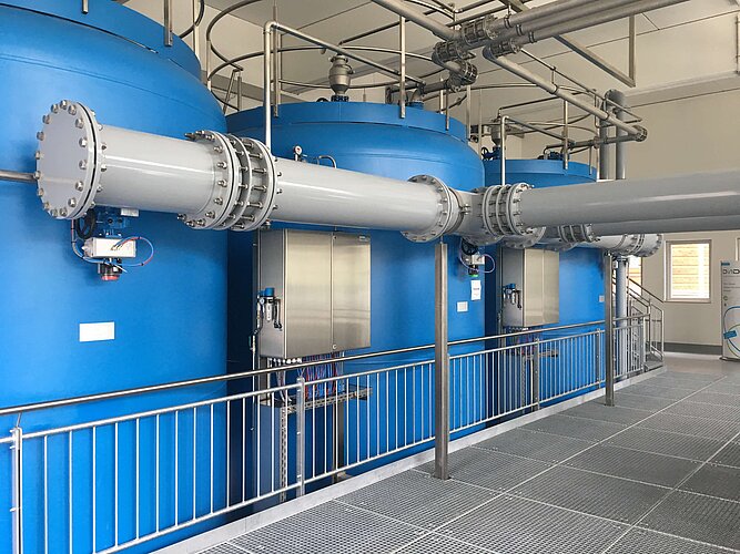 Blaue Tanks mit großen, grauen Rohren verbunden, innerhalb einer Industrieanlage. Die Tanks stehen auf einem Gitterboden und sind von einem Geländer umgeben. Es sind keine spezifischen Maschinen oder Bediener sichtbar.
