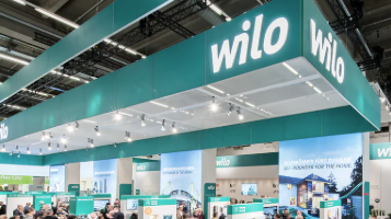 Ein Messestand mit der Aufschrift "Wilo" in großen Buchstaben. Der Stand hat eine moderne, grüne Farbgestaltung und ist mit mehreren Lichtern beleuchtet. Im Hintergrund sind Personen zu sehen, die sich um den Stand versammeln und Informationen einholen.