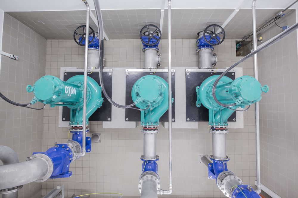 Die Abbildung zeigt eine industrielle Pumpenanlage mit mehreren grünen Pumpen und dazugehörigen Ventilen auf einer grauen Fliesenoberfläche. Es sind Rohre und Verbindungen sichtbar, die die Pumpen miteinander verbinden.