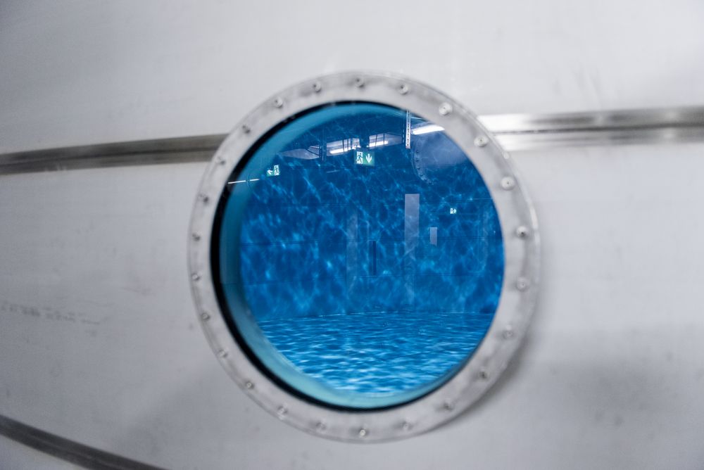 Ein runder Bullaugenfenster zeigt das Wasser eines Schwimmbeckens. Die reflektierenden Wellen und das Licht erzeugen ein sanftes blaues Licht. Im Hintergrund sind schemenhaft Strukturen zu erkennen, die auf eine geschlossene Umgebung hinweisen.