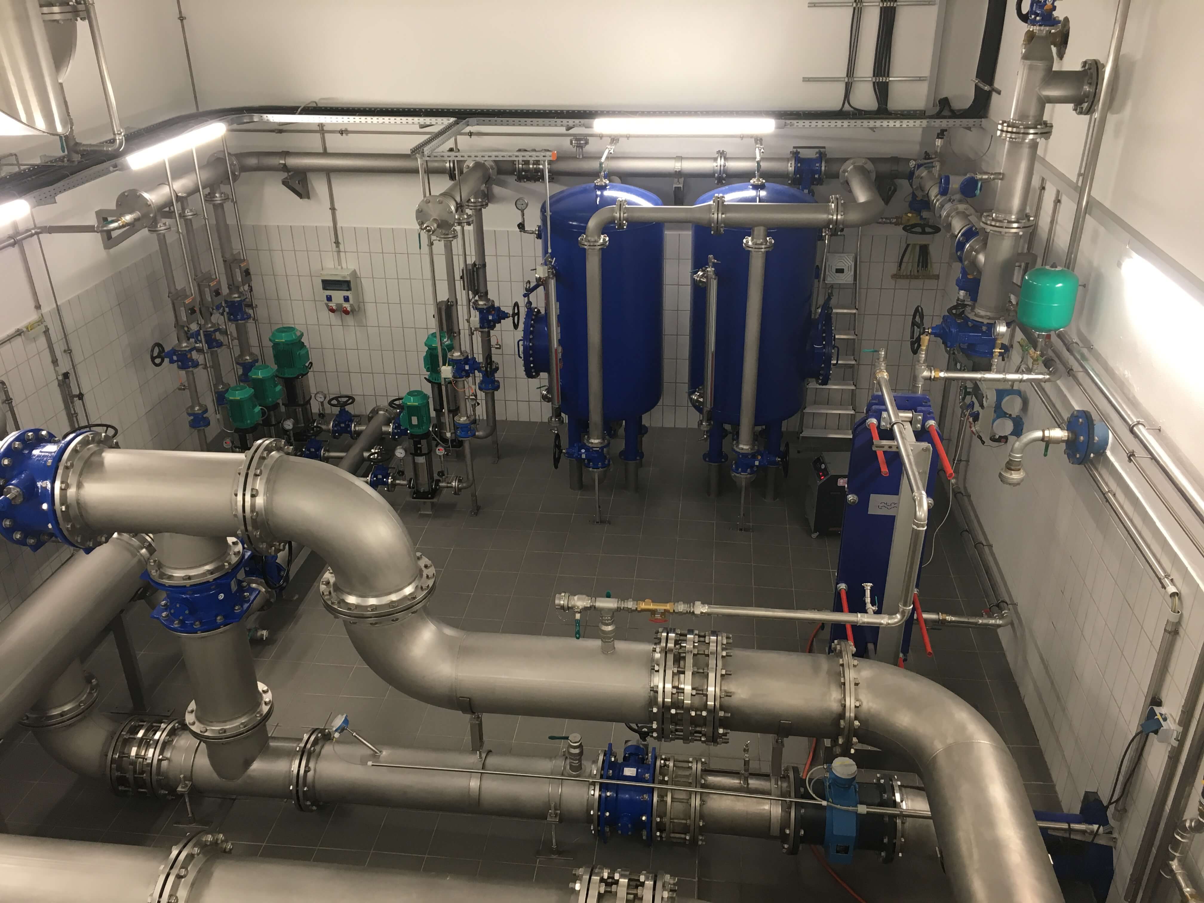 In einem technischen Raum sind große Rohre und zwei große blaue Tanks zu sehen. Die Rohre sind miteinander verbunden und verlaufen durch den Raum. Es gibt mehrere grüne und blaue Geräte, die für einen Wasseraufbereitungsprozess verwendet werden.