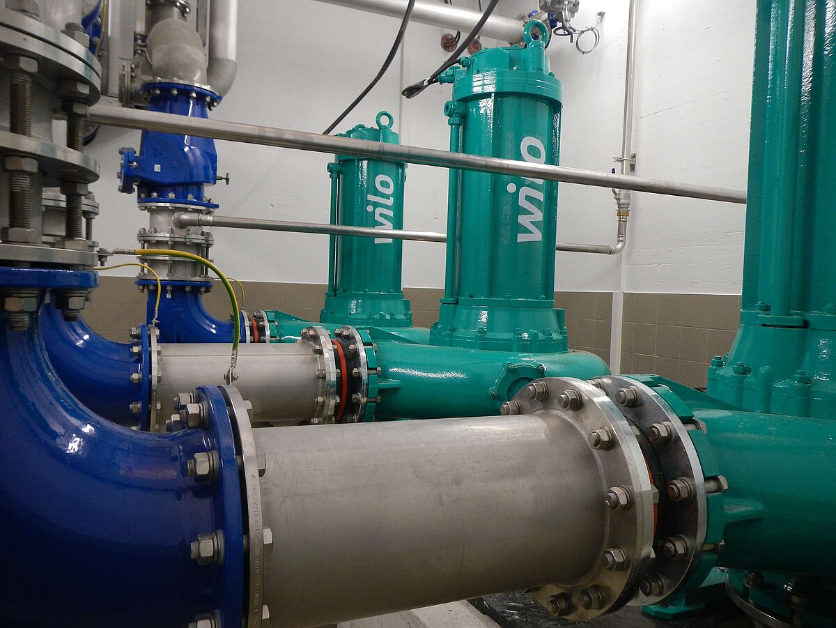 Das Bild zeigt eine industrielle Pumpenanlage mit mehreren grünen Pumpen und blauen Rohren. Die Rohre sind aus Metall und verfügen über Flansche, die mit Schrauben befestigt sind. Die Wand im Hintergrund ist hell und teilweise gefliest.