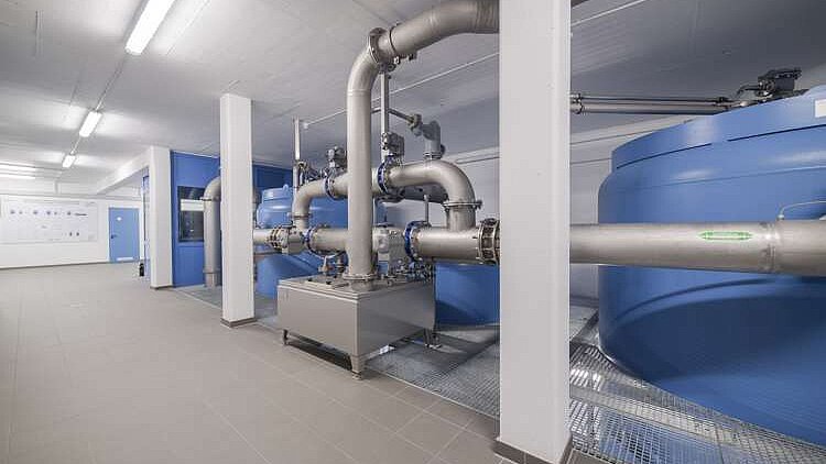 Die Abbildung zeigt einen modernen Raum mit großen blauen Wassertanks und silbernen Rohrleitungen. Die Wände sind in hellen Farben gehalten, und eine Wand zeigt technische Informationen. Der Fußboden ist mit grauen Fliesen ausgelegt und der Raum ist gut beleuchtet.