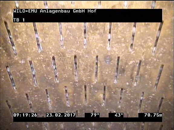 Nahaufnahme einer großen, gestanzten Metalloberfläche mit horizontalen und vertikalen Ausschnitten. Die Oberfläche zeigt Ablagerungen und ist in einem dunklen, industriellen Umfeld. Unterhalb sind technische Informationen und ein Zeitstempel auf einem schwarzen Hintergrund angezeigt.