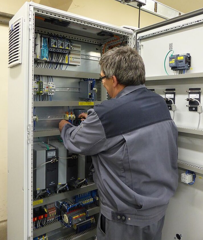 Ein Techniker in grauer Arbeitskleidung arbeitet an einem offenen Schaltkasten, der verschiedene elektronische Komponenten und Kabel zeigt. Der Schaltkasten ist an einer Wand montiert und beleuchtet. Ein Überblick zeigt den Fokus auf die Installation und Wartung von elektrischen Geräten.