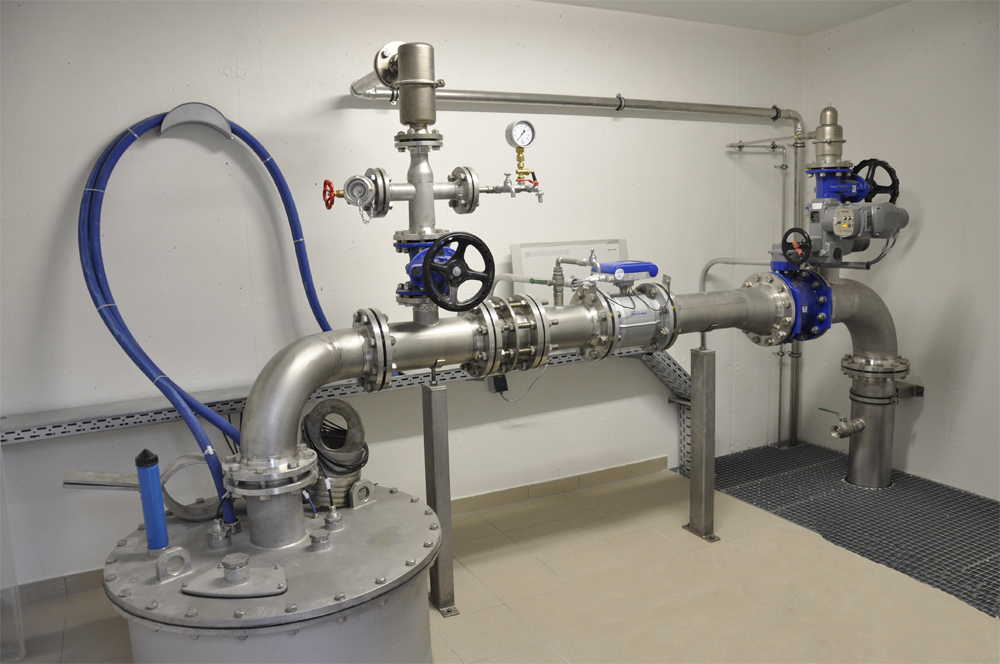 Eine Rohrleitungsanlage in einem Raum mit grauen Wänden. Sie enthält Metallrohre, Ventile und eine Druckanzeige. Die Rohre sind teilweise blau abgedichtet und verlaufen durch den Raum. Der Boden ist mit einem rutschfesten Material ausgelegt.