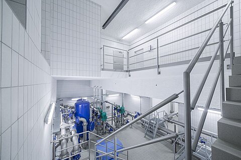 Ein Innenraum eines technischen Gebäudes mit zwei Etagen. Auf der unteren Ebene sind zahlreiche Rohrleitungen, Pumpen und ein großer blauer Tank sichtbar. Eine Treppe führt zur oberen Etage, die ebenfalls mit Geländern und Wänden gefliest ist. Das Licht ist hell und gleichmäßig.