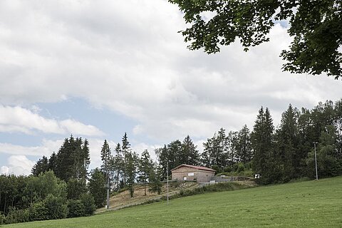 Eine ländliche Szenerie mit einem niedrigen Gebäude auf einem Hügel, umgeben von Bäumen und Wiesen. Der Himmel ist bewölkt, mit einigen aufgelockerten Wolken. Im Vordergrund erstreckt sich eine grüne Wiese.