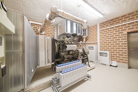 Ein Maschinenraum mit einem großen Motor in der Mitte. Neben dem Motor sind mehrere Batterien angeordnet. Die Wände sind aus rot-ockerfarbenen Ziegeln und eine Metalltür führt zu einem anderen Bereich. Die Decke hat eine helle Beleuchtung.