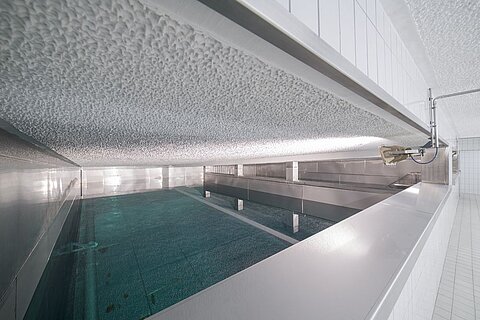 Eine Innenaufnahme eines Schwimmbades mit stilvollem, reflektierendem Wasser und einer modernen, minimalistischen Architektur. Die Wände sind gefliest, und die Beleuchtung ist indirekt, was eine beruhigende Atmosphäre schafft.