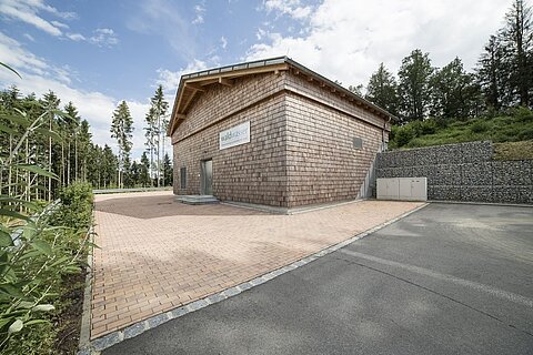 Ein modernes Holzhäuschen mit schrägem Dach steht auf einem gepflasterten Platz, umgeben von Bäumen. Der Eingang ist auf einer Seite sichtbar, während der Platz und die Asphaltstraße davor zu sehen sind. Wolken am Himmel deuten auf einen schönen Tag hin.