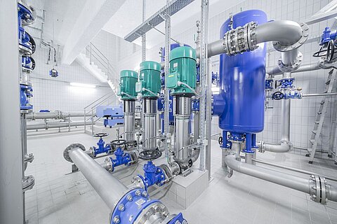 Eine industrielle Pumpenanlage mit grünen und blauen Pumpen, verschiedenen Rohren und Ventilen. Die Wände sind gefliest, und es gibt eine Treppe im Hintergrund. Die gesamte Umgebung ist sehr sauber und gut organisiert.