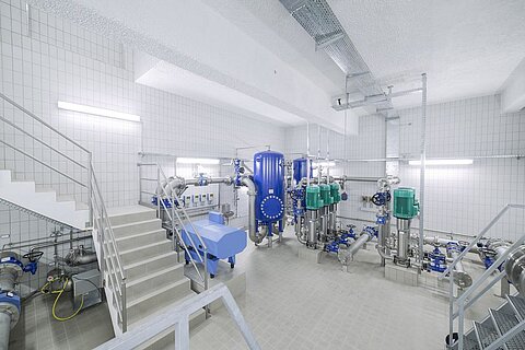 Ein industrieller Raum mit Rohren und Pumpen. Vorne links befindet sich eine Treppe, die in einen anderen Bereich führt. An den Wänden sind elektrische Anlagen und grüne Pumpen zu sehen. Der Raum wirkt sauber und modern, mit hellen Lichtquellen.