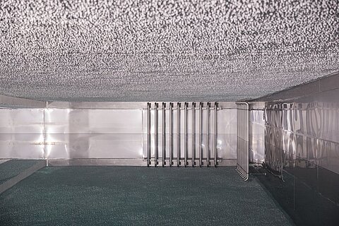 Ein Innenraum mit einem glänzenden metallischen Wandfinish. An einer Wand sind mehrere metallische Rohre angebracht. Der Boden ist mit einer dunklen, strukturierten Oberfläche ausgestattet. Die Decke ist mit einem unregelmäßigen, strukturierten Material versehen.