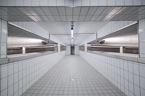 Ein langer, heller Raum mit weißen, glänzenden Fliesen. An den Seiten befinden sich mehrere Arbeitsflächen, und die Decke hat eine gleichmäßige Beleuchtung. Der Boden ist mit quadratischen Fliesen belegt, und es sind keine Möbel oder Dekorationen sichtbar.