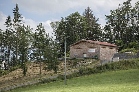 Ein einzelnes, gemauertes Gebäude mit rotem Dach steht auf einem Hügel, umgeben von Bäumen und Wiese. Ein Gehweg führt zum Gebäude, das in einer ländlichen Umgebung gelegen ist. Der Himmel ist bewölkt.