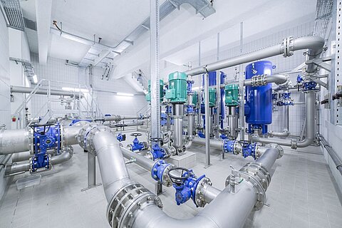 Ein Raum mit umfangreichen Rohrleitungen und Ventilen. Mehrere große blauen und grünen Behälter sind an den Rohren befestigt. Die Wände sind hell gefliest, und die Beleuchtung ist gleichmäßig, was auf eine industrielle Umgebung hinweist.