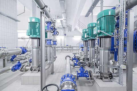 Industriehalle mit Rohrleitungssystemen und mehreren Wasserpumpen in grüner und blauer Farbgebung. Die Umgebung ist hell und modern, mit sichtbaren Rohren, Ventilen und Regalflächen im Hintergrund.