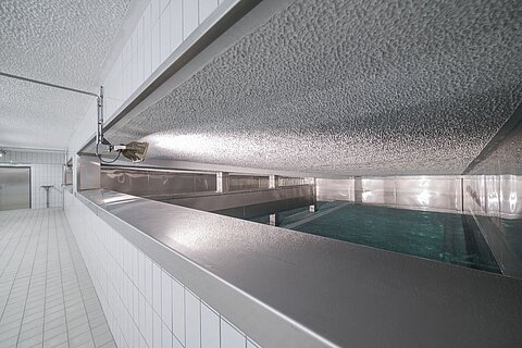 Ein Innenraum mit einem langen, schmalen Wasserbecken aus Edelstahl. Die Wände sind gefliest und die Beleuchtung ist weich. Es gibt eine Wandöffnung und eine Rohrleitung, die von der Decke hängt, aber keine Menschen oder andere Gegenstände sind sichtbar.