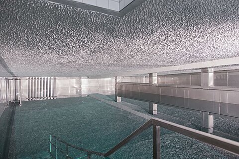 Eine moderne Innenarchitektur mit einem glatten, reflektierenden Boden und einer gewölbten Decke. Das Licht fällt gleichmäßig auf die Fläche und erzeugt eine ruhige Atmosphäre. Es gibt eine geschwungene Treppe, die zu einem oberen Bereich führt.