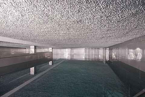 Der Raum zeigt eine strukturierte Decke mit glatten Wänden und einem schimmernden Boden. Es gibt glatte Oberflächen und eine Reihe von metallischen Rohren, die die Wände entlang verlaufen. Die Beleuchtung erzeugt ein modernes und kühles Ambiente.