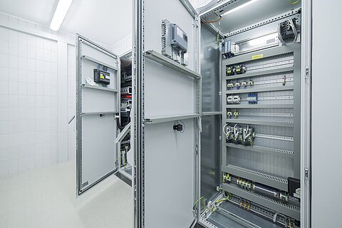 Blick in einen technischen Raum mit geöffneten Schaltschränken. Die Schaltschränke sind mit verschiedenen elektrischen Komponenten und Schaltkreisen ausgestattet. Der Raum hat eine helle, gepflegte Atmosphäre mit weißen Fliesen und Deckenbeleuchtung.