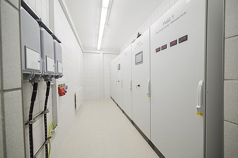Ein schmaler Raum mit weißen Wänden und einem hellen Boden. Links sind mehrere elektrische Schaltschränke angebracht, mit digitalen Anzeigen und Schaltern. Rechts befindet sich eine geschlossene Tür zu einem weiteren Bereich. Die Beleuchtung ist gleichmäßig und hell.