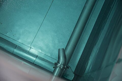 Eine Nahaufnahme von einem metallischen Rohr, das an einer Wand befestigt ist. Die Umgebung wirkt kühl und reflektiert einige Lichtreflexe. Der Hintergrund zeigt eine glatte, teilweise spiegelnde Oberfläche.