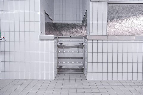 Ein sauberer Raum mit weißen Fliesen, der eine Öffnung in einer Wand zeigt, durch die ein metallisches Regal sichtbar ist. Die Wände sind glatt und reflektierend, und der Boden ist mit grauen Fliesen ausgelegt.