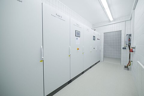 Ein Raum mit hellen, weißen Wänden und einem glatten, hellen Boden. An der linken Wand stehen mehrere große, geschlossene Schränke mit Bedienfeldern. Im Hintergrund ist ein Türöffner zu einem weiteren Raum sichtbar.