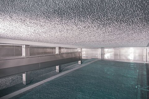 Ein moderner, leerer Raum mit einer reflektierenden, glatten Oberfläche. Die Wände und die Decke sind aus Metall und scheinen mit einer glänzenden Schicht überzogen zu sein. Der Boden ist mit Wasser gefüllt, das eine ruhige, blaue Farbgebung hat.