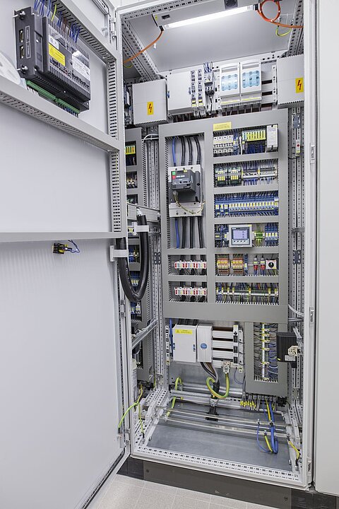 Ein geöffneter Schaltkasten, der mit verschiedenen elektrischen Komponenten, Kabeln und Steuerungen ausgestattet ist. Die Innenseite ist beleuchtet und zeigt eine ordentliche Anordnung von Leitungen und Geräten für die elektrische Steuerung.