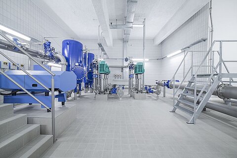 Ein moderner Maschinenraum mit blauen Rohren und Pumpen. Sichtbare Treppen führen zu einer höheren Plattform. Der Boden ist gefliesst und es gibt mehrere technische Geräte und Steuerkästen an den Wänden. Helle Beleuchtung sorgt für eine klare Sicht auf die Ausrüstung.