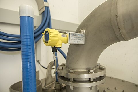 Ein gelbes Messgerät ist an einem grauen Rohr angebracht, das Teil eines industriellen Systems ist. Neben dem Rohr befindet sich ein blauer Schlauch. Im Hintergrund sind weitere Rohre und Kabel sichtbar.