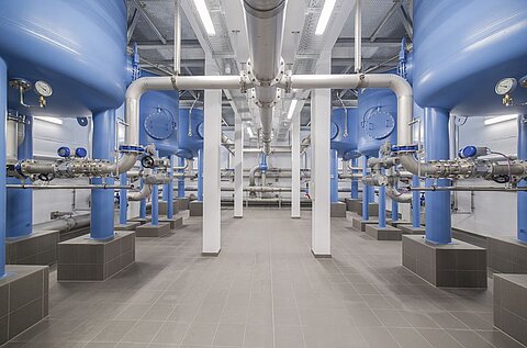 Innenansicht einer modernen Industrieanlage mit blauen Wassertanks, Rohrleitungen und Instrumenten zur Messung. Die Wassertanks stehen auf blauen Säulen, umgeben von grauen Fliesen. Das Licht ist hell und gleichmäßig verteilt.