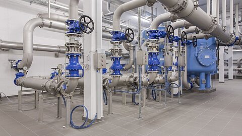 Das Bild zeigt eine industrielle Rohrleitungsanlage mit mehreren Rohren und Ventilen. Die Rohre sind aus Metall, während einige Ventile und Komponenten blau gefärbt sind. Die Anordnung ist ordentlich und modern, typisch für industrielle Anwendungen in der Wasser- oder Abwassertechnik.