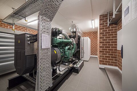 Eine Gruppe von Maschinen in einem Raum, darunter ein großer grüner Motor und verschiedene technische Geräte. Die Wände sind mit roten Ziegeln verkleidet, und es gibt Regale sowie einen Schrank im Hintergrund. Deckenleuchten sorgen für Helligkeit.