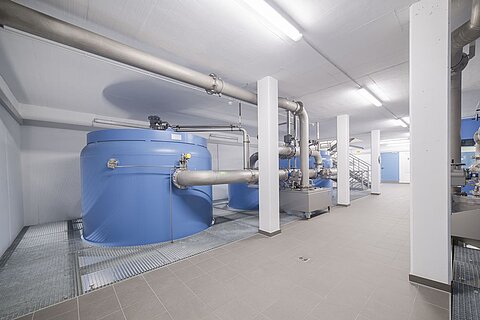 Innenraum einer Industrieanlage mit blauen Wassertanks und mehreren Rohrleitungen. Der Boden ist gefliest, und es gibt eine Treppe im Hintergrund. Die Lichtverhältnisse sind hell und gleichmäßig.