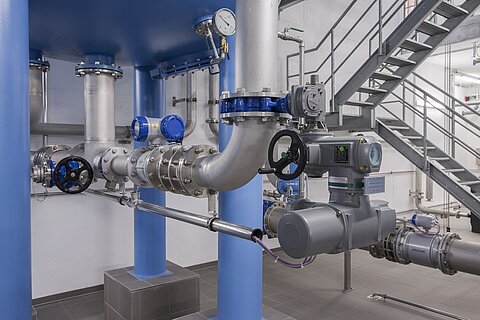 Das Bild zeigt ein industrielles Rohrleitungssystem mit metallischen Rohren, Ventilen und Anzeigegeräten. Im Hintergrund befindet sich eine Treppe. Die Leitungen sind farblich markiert und deutlich sichtbar, was auf eine technische Umgebung hinweist.