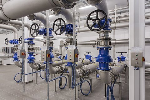 Eine industrielle Rohrleitungsanlage mit mehreren blau lackierten Absperrhähnen und Rohren. Die Struktur besteht aus mehreren Rohren, Ventilen und Steuerpanelen. Die Umgebung ist sauber und modern, mit grauen Fliesen und weißen Wänden.