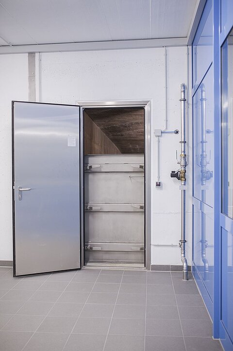 Ein offenes, metallisches Türblatt zu einem Kühlraum oder einer Kühlzelle in einem modernen Raum. Die Wände sind hell und die Fliesen sind grau. Neben der Tür sind Rohrleitungen sichtbar.