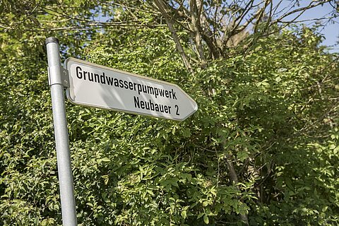 Ein Verkehrsschild, das auf das Grundwasserpumpwerk und die Neubauer Straße 2 hinweist, steht vor dichtem, grünem Laub.
