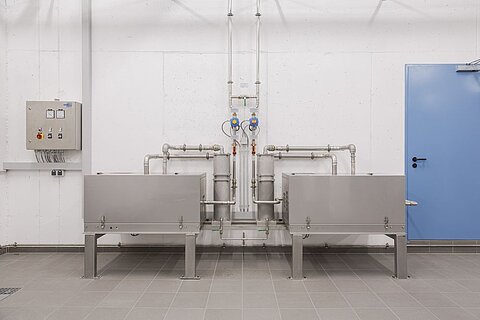 In einem modernen Raum sind zwei graue Behälter auf Metallfüßen aufgestellt, verbunden durch Rohre. An der Wand befindet sich ein Schaltkasten mit Tasten und Anzeigen. Eine Tür in Blau ist ebenfalls sichtbar.