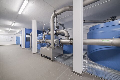 In einem modernen Raum sind große blaue Wassertanks und dazugehörige Rohrleitungen zu sehen. Der Boden ist mit Fliesen ausgestattet, und die Wände sind hell gestrichen. Es gibt auch eine graue Plattform, auf der die Rohrsysteme montiert sind.