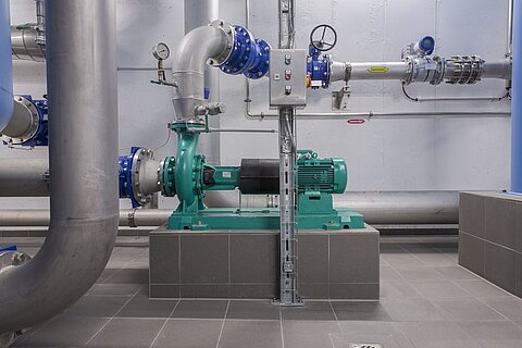 Eine grüne Pumpe mit Elektromotor steht auf einem grauen Podest, umgeben von großen Metallrohren. Die Pumpe ist Teil eines technischen Systems in einem industriellen Raum, der mit grauen Fliesen ausgelegt ist.
