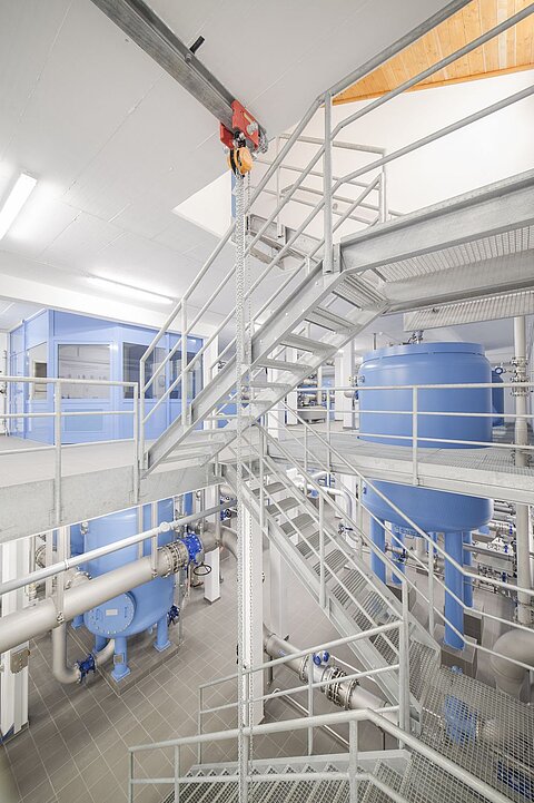 Im Bild ist eine industrielle Umgebung mit einer metallenen Treppe zu sehen, die in einem Raum mit blauen Tanks und Rohren führt. Der Boden ist gefliest, und es gibt verschiedene Maschinen und technische Einrichtungen im Hintergrund.