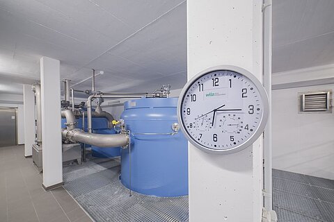 Ein Innenraum mit einem großen blauen Wasserbehälter und Rohren. An der Wand hängt eine analoge Uhr, die die Zeit anzeigt. Der Boden besteht aus grauen Fliesen und die Wände sind hell. Es ist gut beleuchtet.