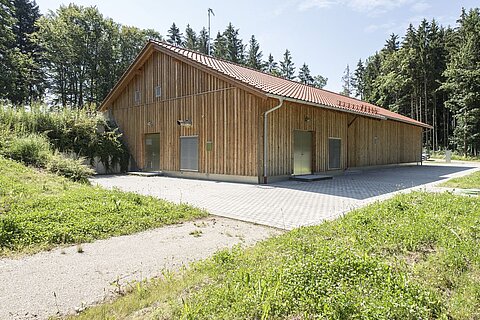Ein modernes, zweigeschossiges Gebäude aus Holz mit einem roten Satteldach, umgeben von grünen Wiesen und Bäumen. Der Eingang ist seitlich und der Boden ist gepflastert.